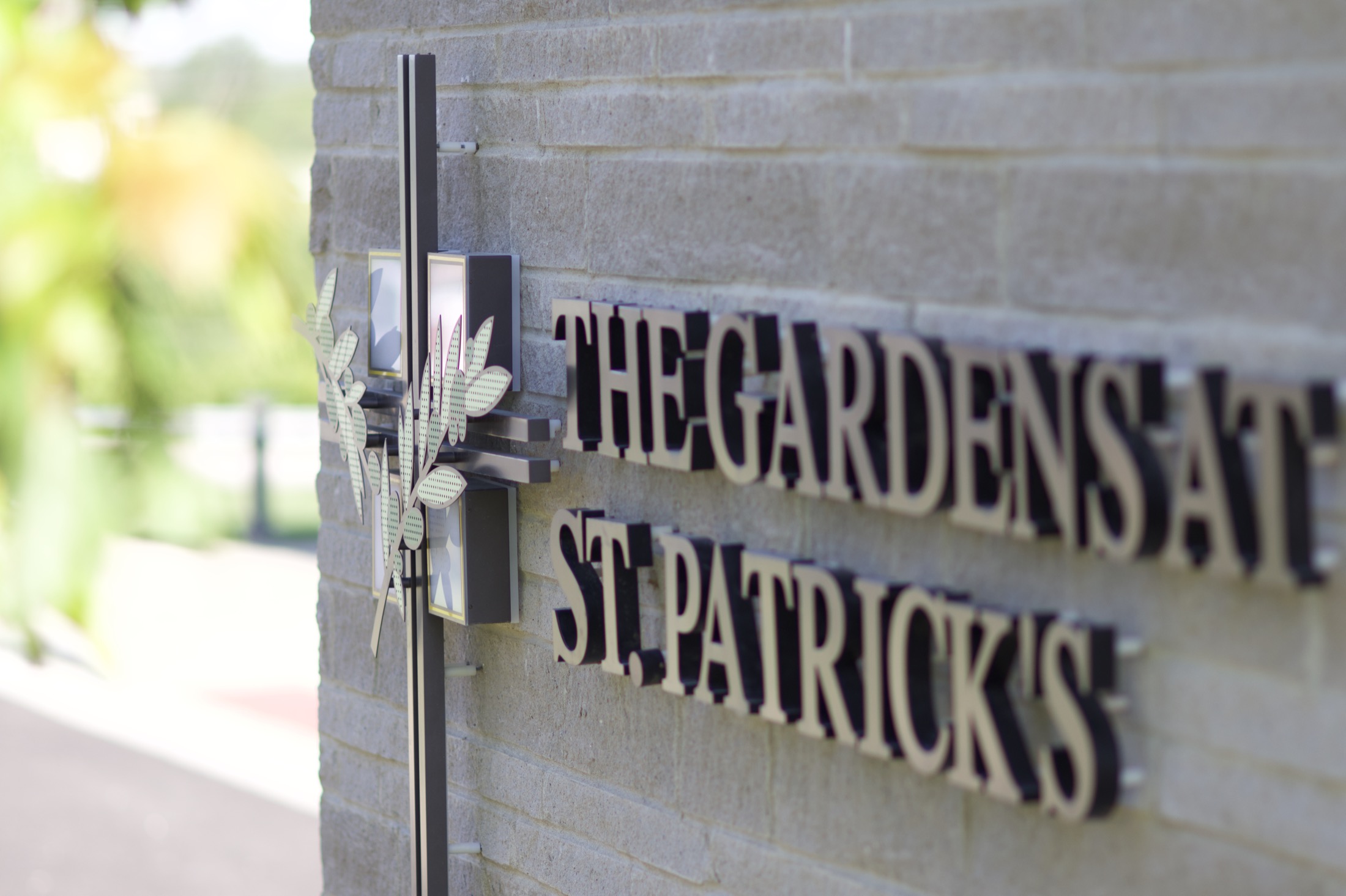 A carved stone sign reading The Gardens at St. Patrick's, mounted beside an olive branch bracket at the entrance.