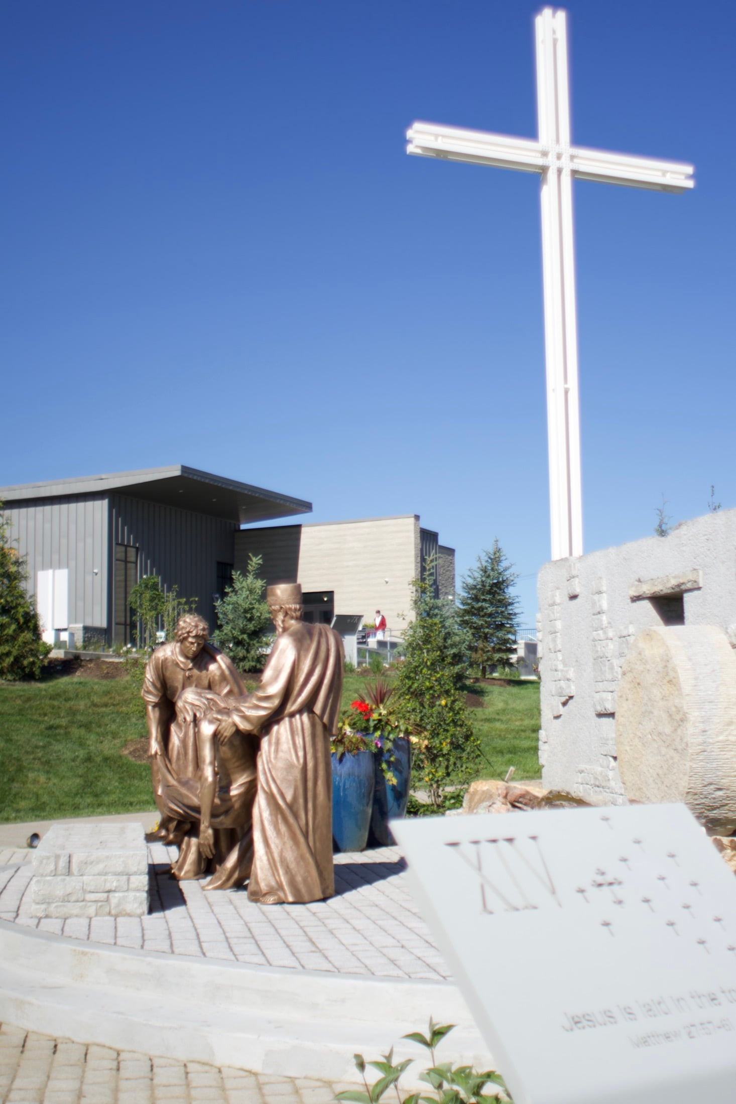 A cross rising against the sky beside a station marker inscribed with raised braille, the Welcome Center beyond.