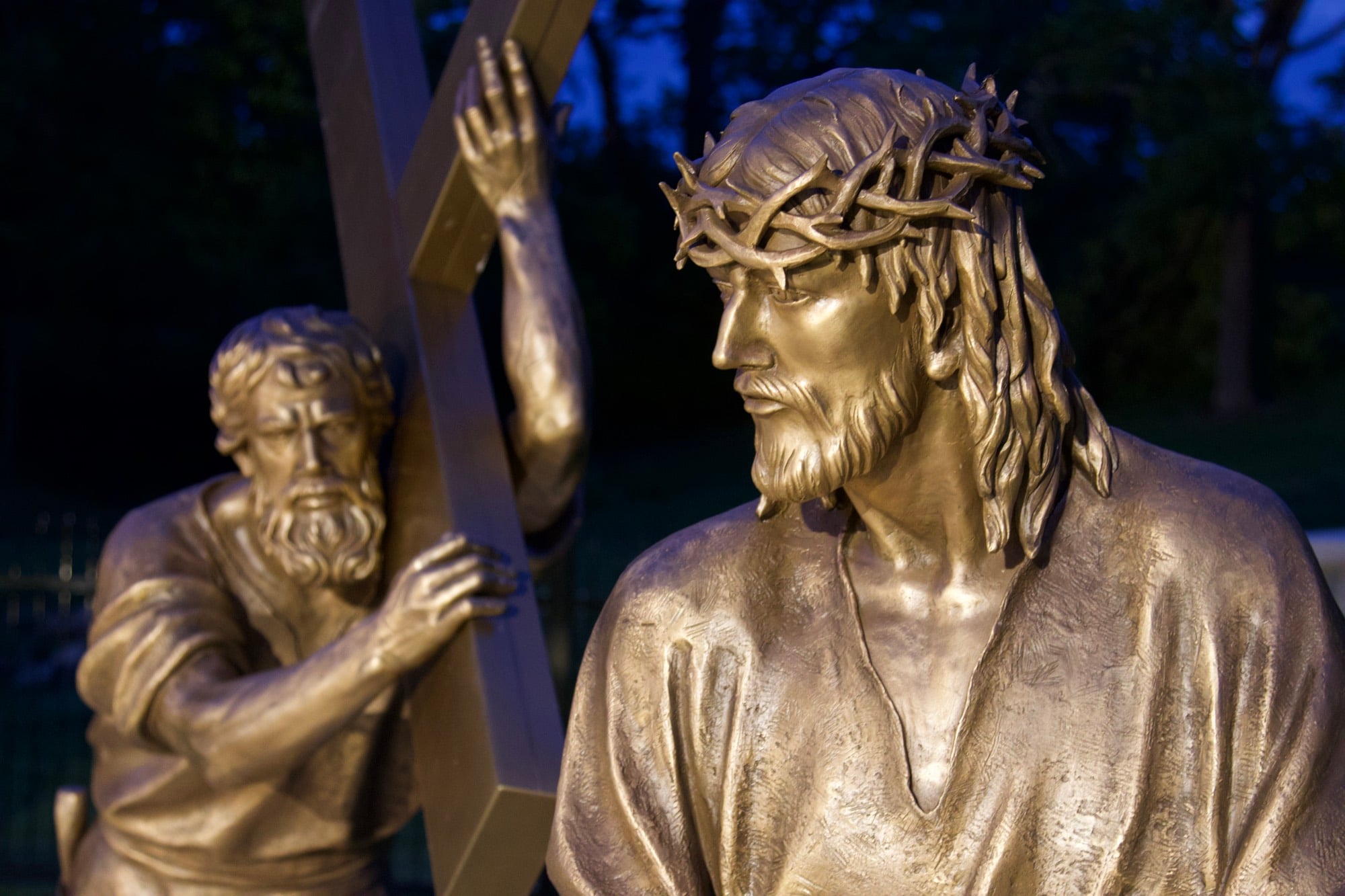 Bronze figures of Christ and Simon of Cyrene lit against a deep blue evening sky.