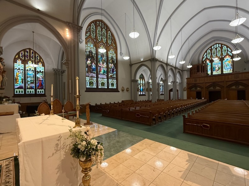 View across the nave from the altar, showing the ribbed vaulted ceiling, tall stained-glass windows, and rows of wooden pews.