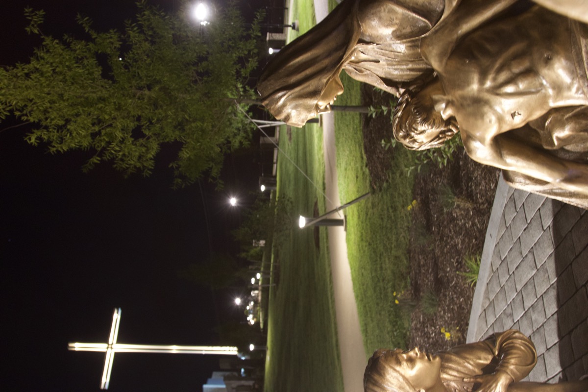 A bronze Pietà beside the lit path, the monumental cross glowing down the garden at night.