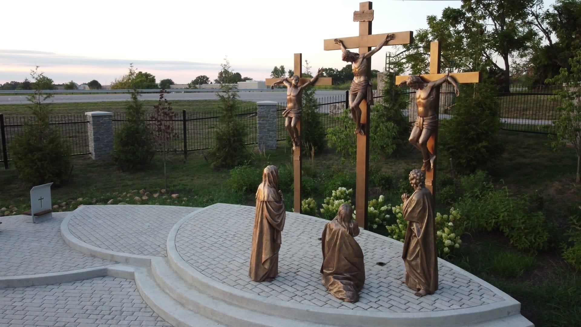 Aerial view of the bronze Crucifixion: Christ on the central cross flanked by the two thieves, with mourning figures at the base, at dusk.